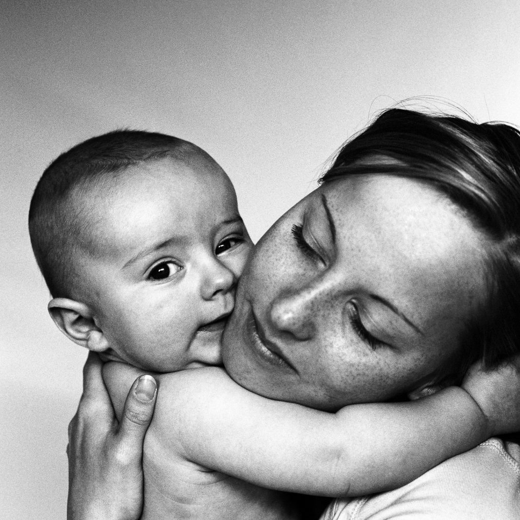 Baby kuschelt mit Mama und blickt beim Baby Fotoshooting zuhause in die Kamera