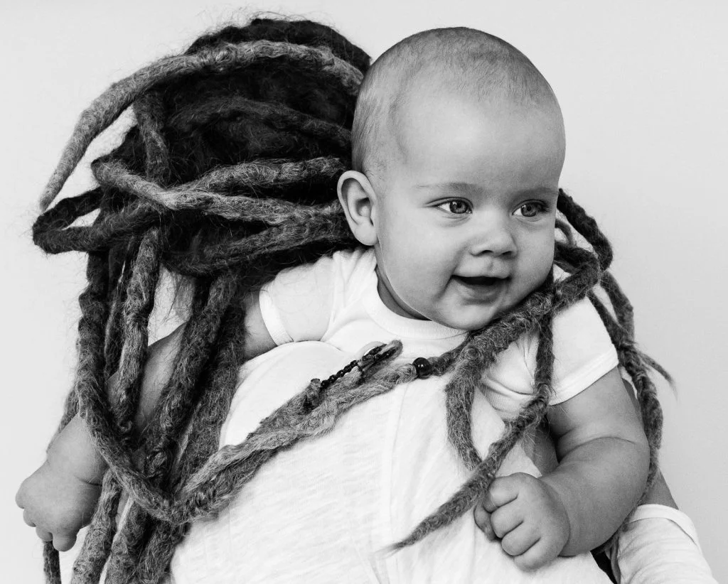 Baby schaut über die Schulter der Mama und ist umgeben von Rastazöpfen beim Baby Fotoshooting in Hamburg Eppendorf