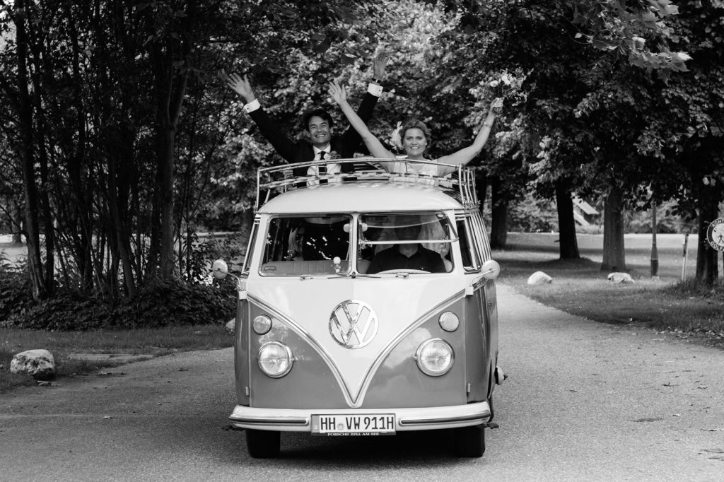 Das Brautpaar winkt aus dem Hochzeitsauto VW Bus T1 am Goldschätzchen der Koch Fabrik auf dem Golf Park Peiner Hof in Priesdorf beim Hochzeitsfotograf Hamburg