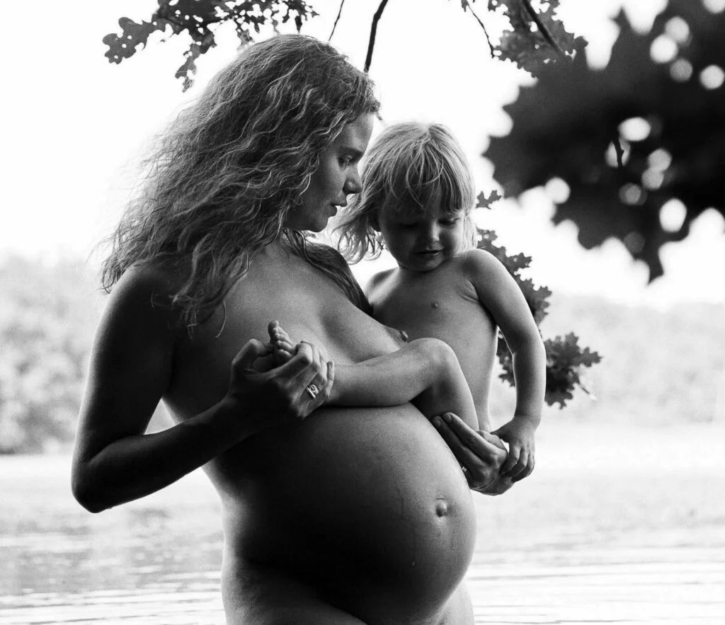 Schwangere steht im Wasser und hält ihr Kleinkind auf dem Arm. Gemeinsam schauen sie auf den Babybauch bei den Babybauchfotos in Hamburg in der Natur am See