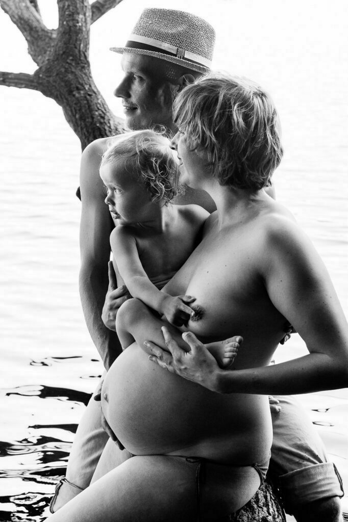 Mama und Papa sitzen gemeinsam mit ihrem Kind im Arm am See und schauen auf das Wasser bei den Babybauchfotos in Hamburg in der Natur