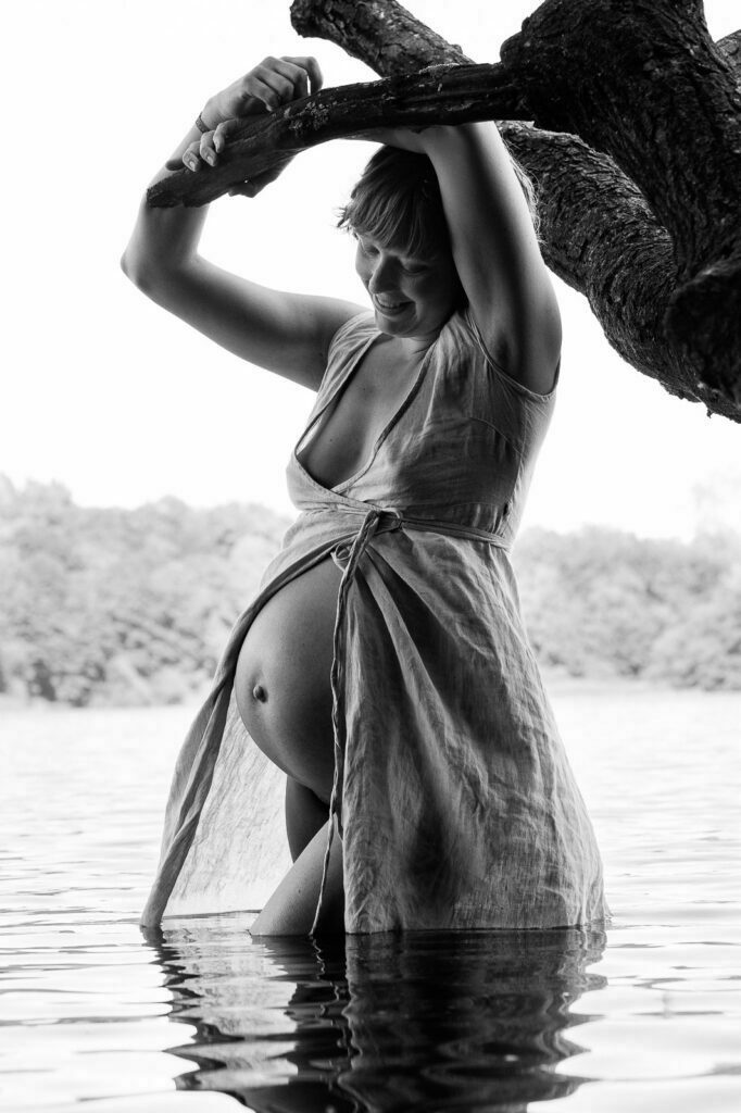 Schwangere steht im Kleid im Wasser und schaut auf ihren Babybauch bei den Babybauchfotos in Hamburg am See in der Natur