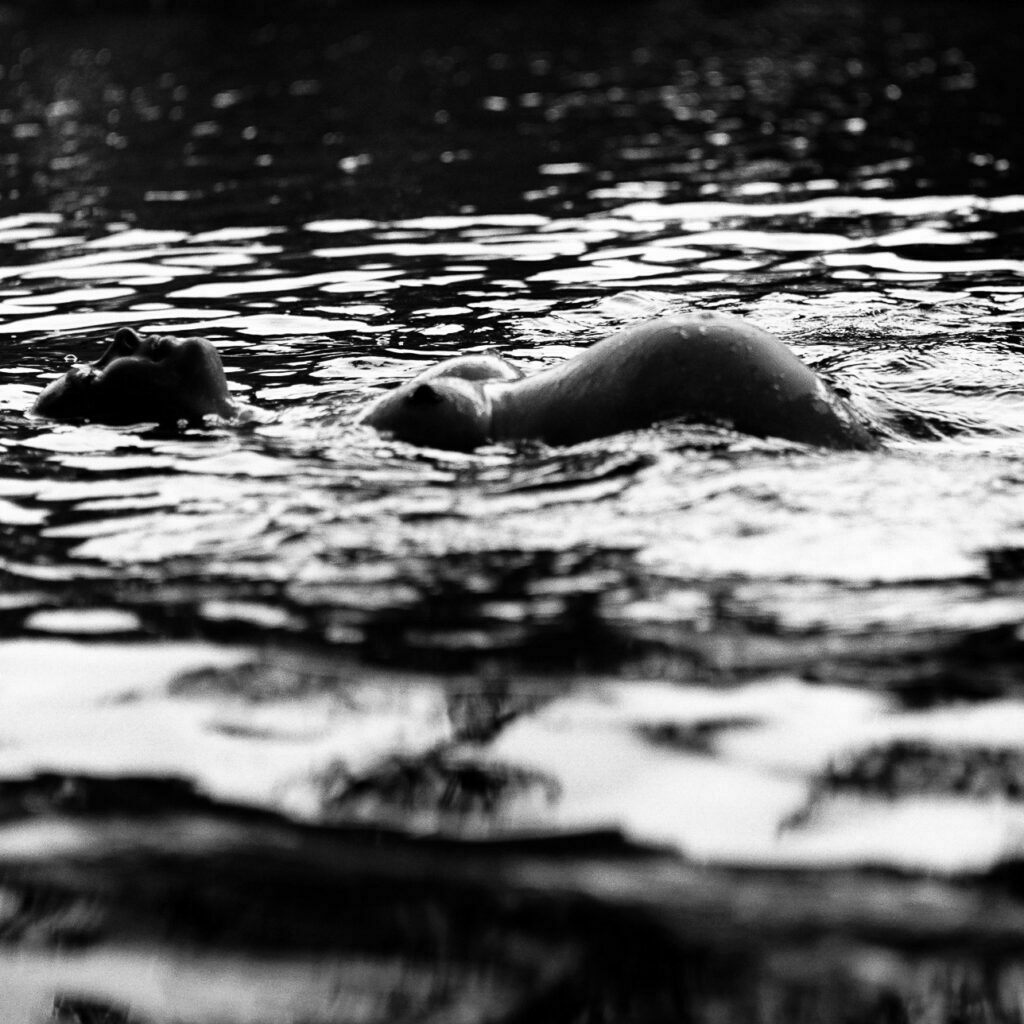 Schwangere schwimmt auf dem Rücken im See und hebt dabei ihren Babybauch aus dem Wasser bei den Babybauchfotos in Hamburg in der Natur